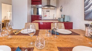 Uma sala de jantar com mesa de madeira posta para quatro, cozinha ao fundo e relógio na parede.