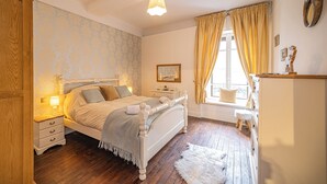 Quarto com cama, mesas de cabeceira, cômoda, janela com cortinas e radiador.