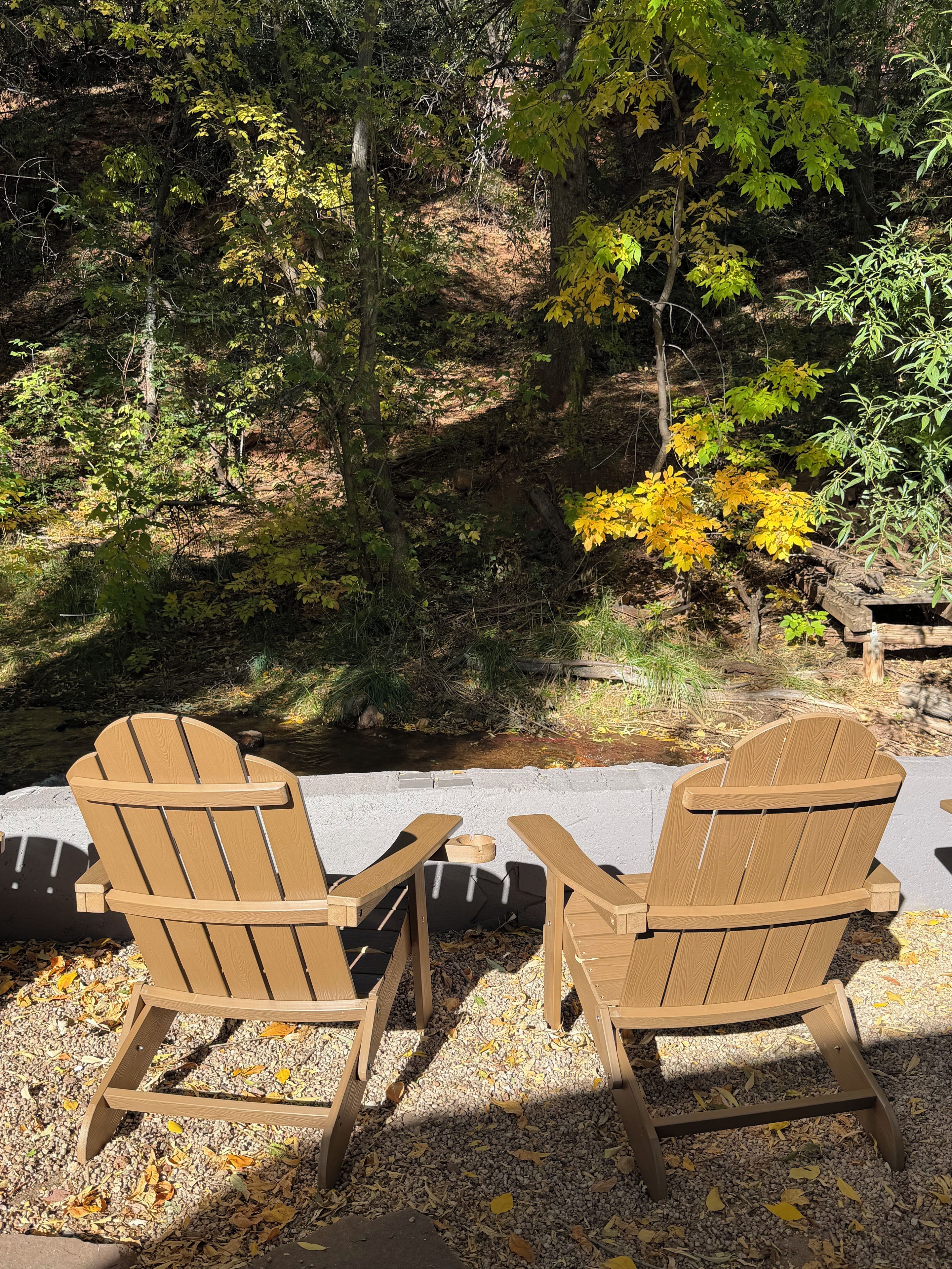 Two wooden Adirondack chairs facing each other with a concrete ledge in between, set against a backdrop of trees and a small stream.