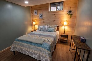 Ein Schlafzimmer mit einem hölzernen Kopfteil, zwei Nachttischlampen, einem Bett mit gemusterten Bettwaren, einem kleinen Tisch und einem Fenster mit Aussicht.