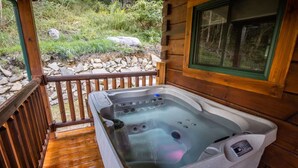 Un jacuzzi en una terraza de madera con un muro de piedra y follaje al fondo.