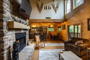 Ein gemütliches Blockhaus-Interieur mit Kamin aus Stein, Essbereich aus Holz und einem großen Fenster mit Blick ins Freie.