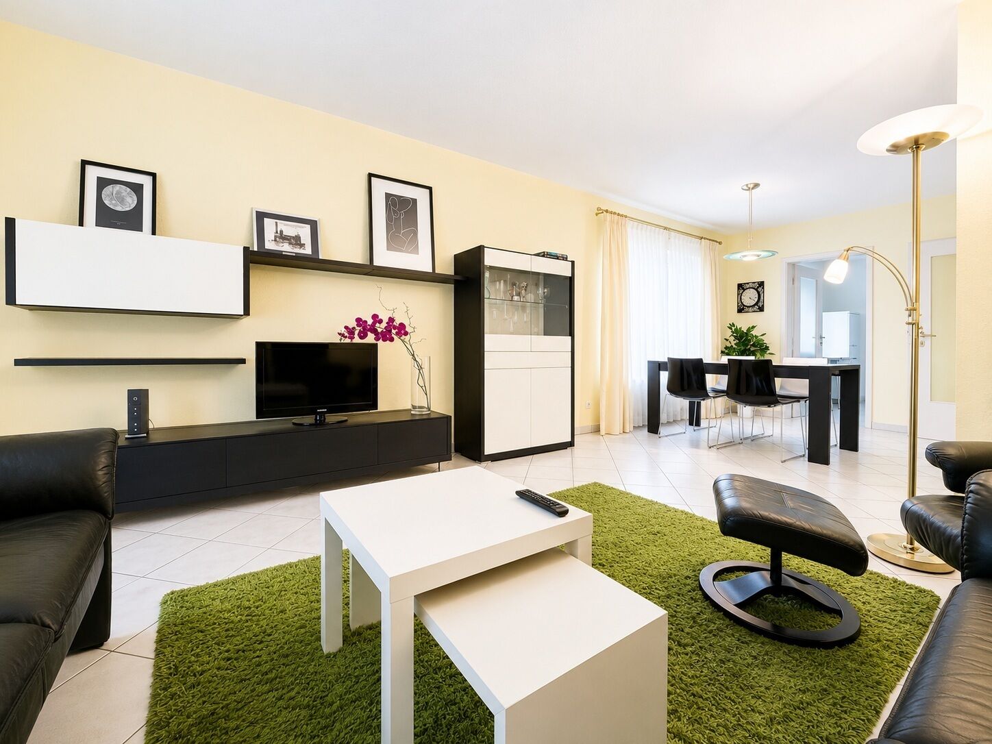 Ein modernes Wohnzimmer mit Flachbildfernseher, Couchtisch, Sofa, Essbereich mit Tisch und Stühlen sowie Küchenzeile mit Schränken und Kühlschrank.