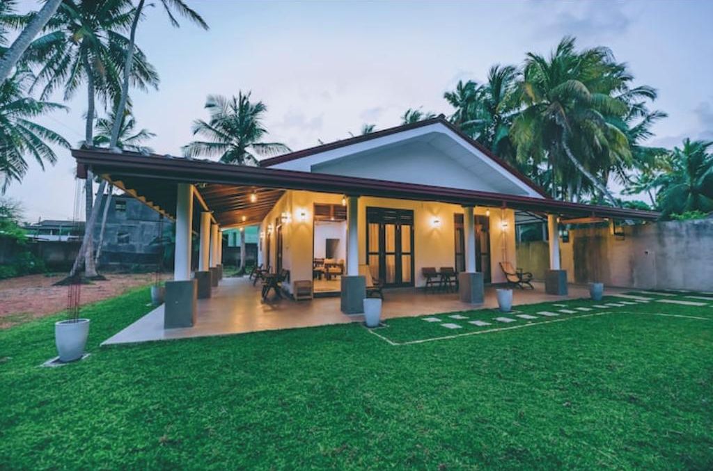 Uma casa moderna com um alpendre coberto, gramado e palmeiras ao fundo.