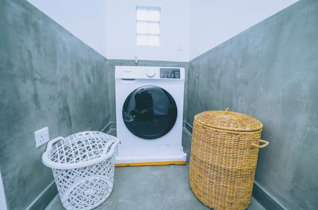 Un cuarto de lavandería con una lavadora, un cesto blanco para ropa y un cesto de mimbre.