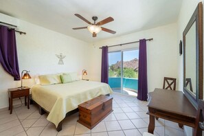 Quarto com cama, luminárias de cabeceira, ventilador de teto, um baú de madeira, escrivaninha e vista para uma piscina e montanhas por uma porta de correr.