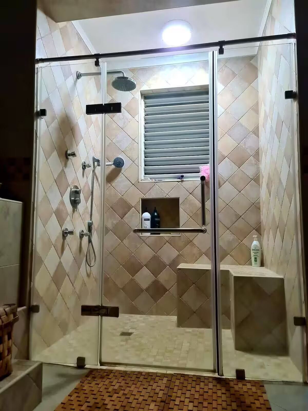 Et bruseområde med glasvægge, en flisebelagt væg, et vindue med skodder og en hylde med toiletartikler.