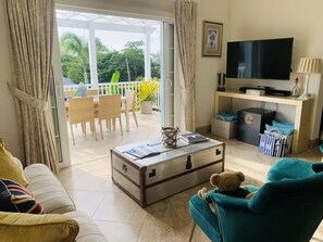 A living room with a sofa, a teal armchair, a coffee table with a suitcase, a television, and a balcony with outdoor furniture. - Your Private Luxury Caribbean Retreat In Barbados Pool Sea Views -walk to beach! (Gibbes)