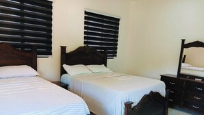 Habitación de hotel con dos camas, cabecera de madera, mesita de noche, cómoda y ventana con persianas.