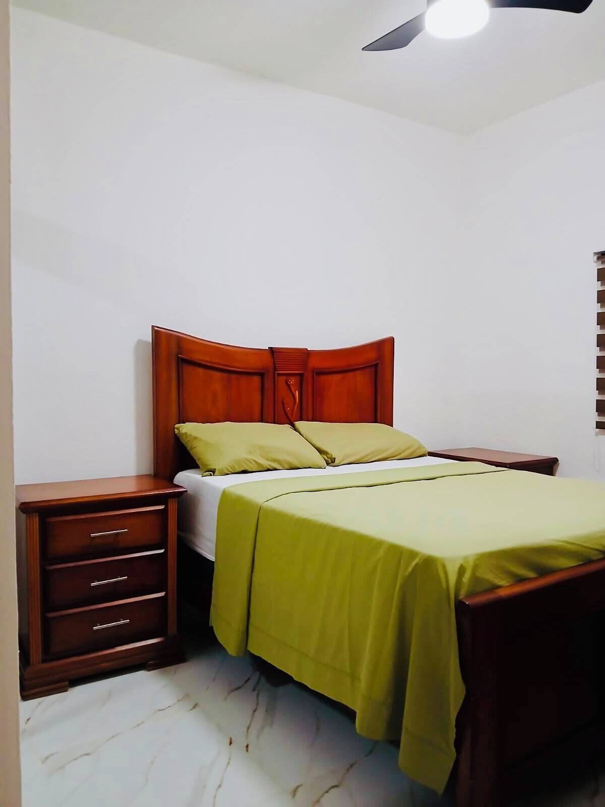 Ein Schlafzimmer mit einem hölzernen Bettgestell, einer grünen Bettdecke, einem Nachttisch mit Schubladen und einem Deckenventilator.