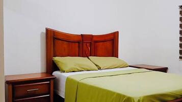 Un dormitorio con estructura de cama de madera, colcha verde, mesita de noche con cajones y ventilador de techo.