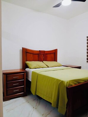 A bedroom with a wooden bed frame, a green bedspread, a nightstand with drawers, and a ceiling fan.