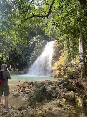 A waterfall in a forested area with a person taking a photo. - Santa Capuza Las Brisas (Sánchez)