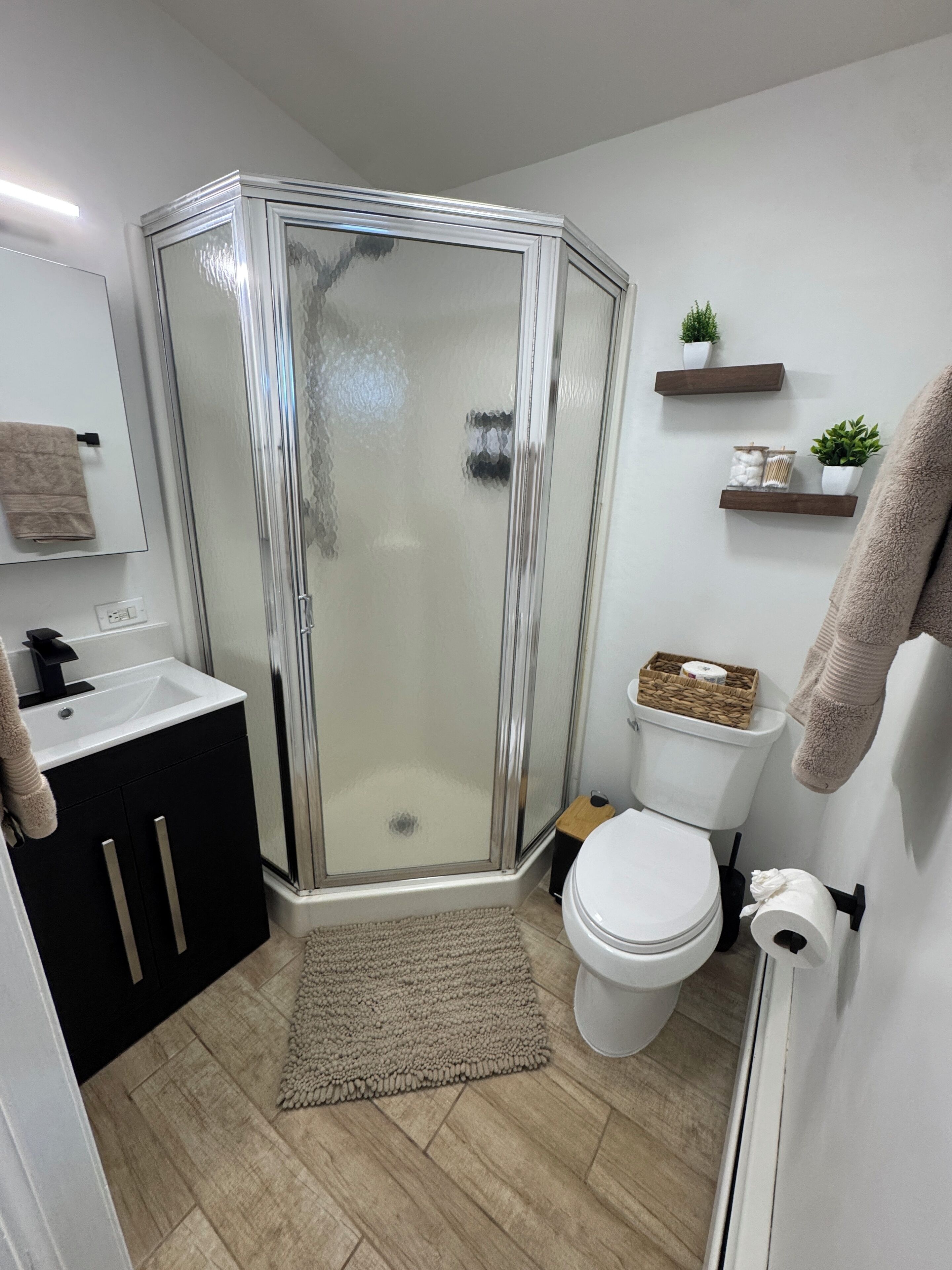 Ein modernes Badezimmer mit Duschabtrennung aus Glas, einer weißen Toilette, einem Waschbecken mit schwarzem Unterschrank und einem beigen Teppich.