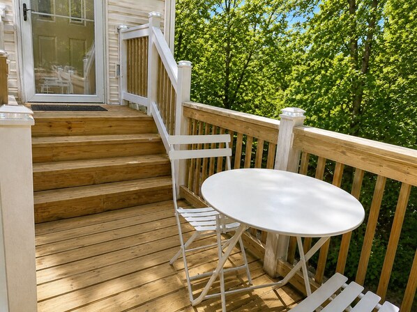 A wooden deck with a white table and chairs, a glass door, and a view of green trees.