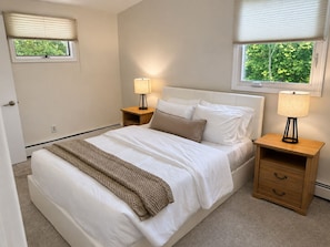 A bedroom with a bed, two bedside tables, a window with blinds, and a view of greenery outside.