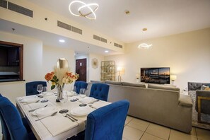 A dining area with a table set for six, blue upholstered chairs, a vase with flowers, and a television displaying a welcome message. - 3BR+Maid in JBR | Near The Walk & JBR Beach (Dubai)
