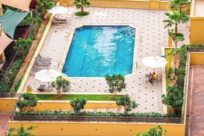 Ein Swimmingpool mit Liegestühlen und Sonnenschirmen, umgeben von viel Grün und einer gefliesten Terrasse.