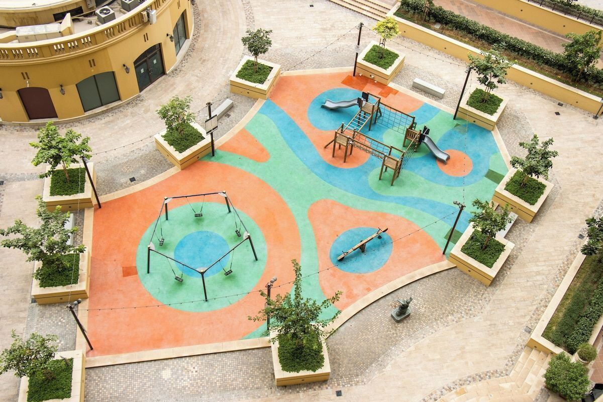 A playground with swings, a slide, and climbing structures surrounded by planters with greenery.