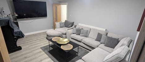 A living room with a grey sofa set, a flat-screen TV mounted on the wall, and a coffee table with decorative items.