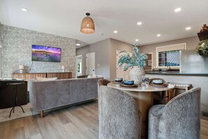Un salon moderne avec un canapé gris, une table à manger avec des chaises, un luminaire suspendu et un téléviseur fixé au mur.