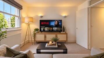 A living room with a television displaying Netflix shows, a lamp, a bookshelf, and a coffee table with books and a candle.