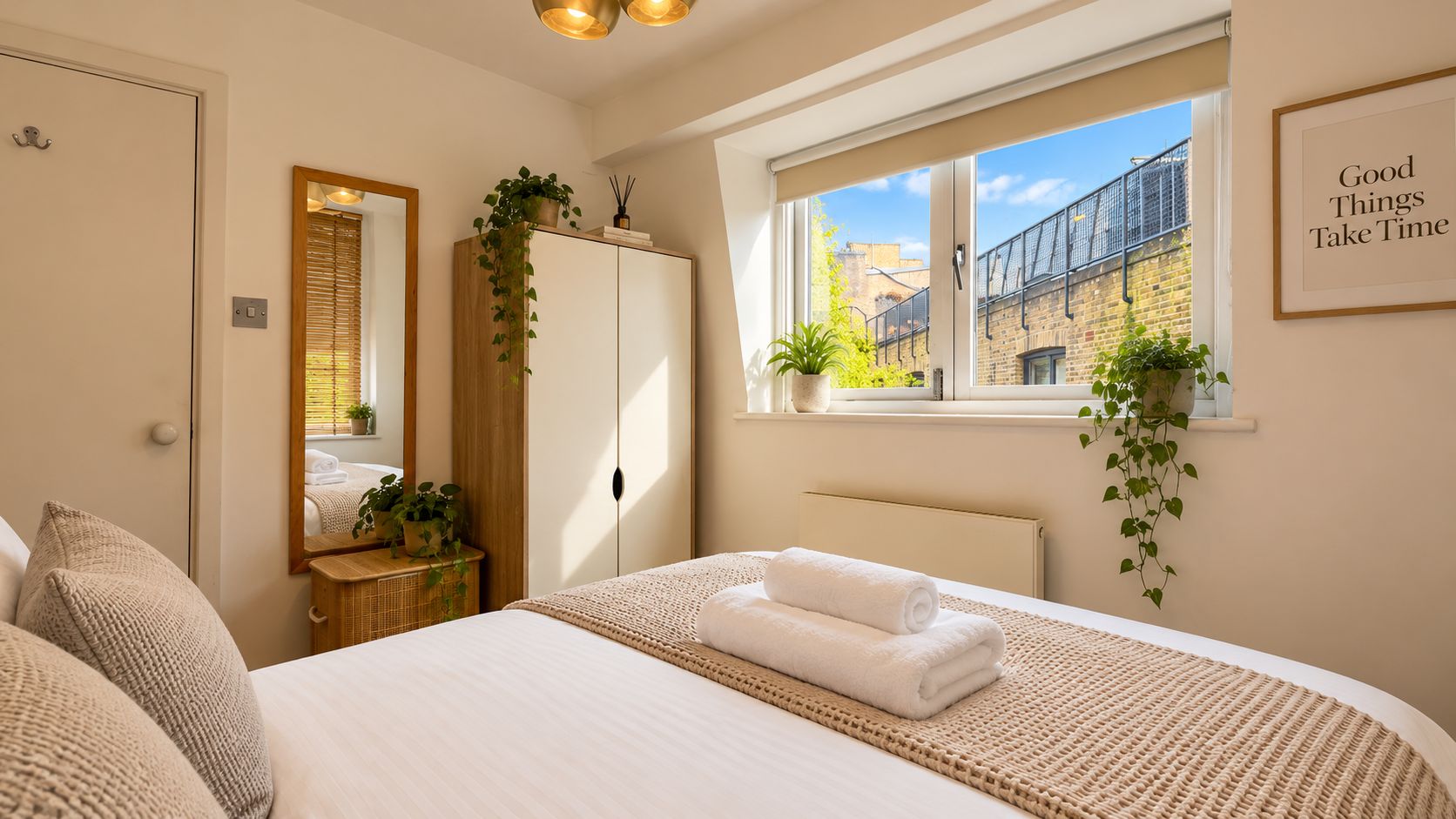A bedroom with a bed, a wardrobe, a bedside table with a plant, a window with a view of buildings, and a motivational quote on the wall.