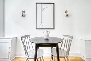 Een eetgedeelte met een ronde tafel, twee stoelen, een witte vaas en een lijst met abstracte kunst aan de muur.