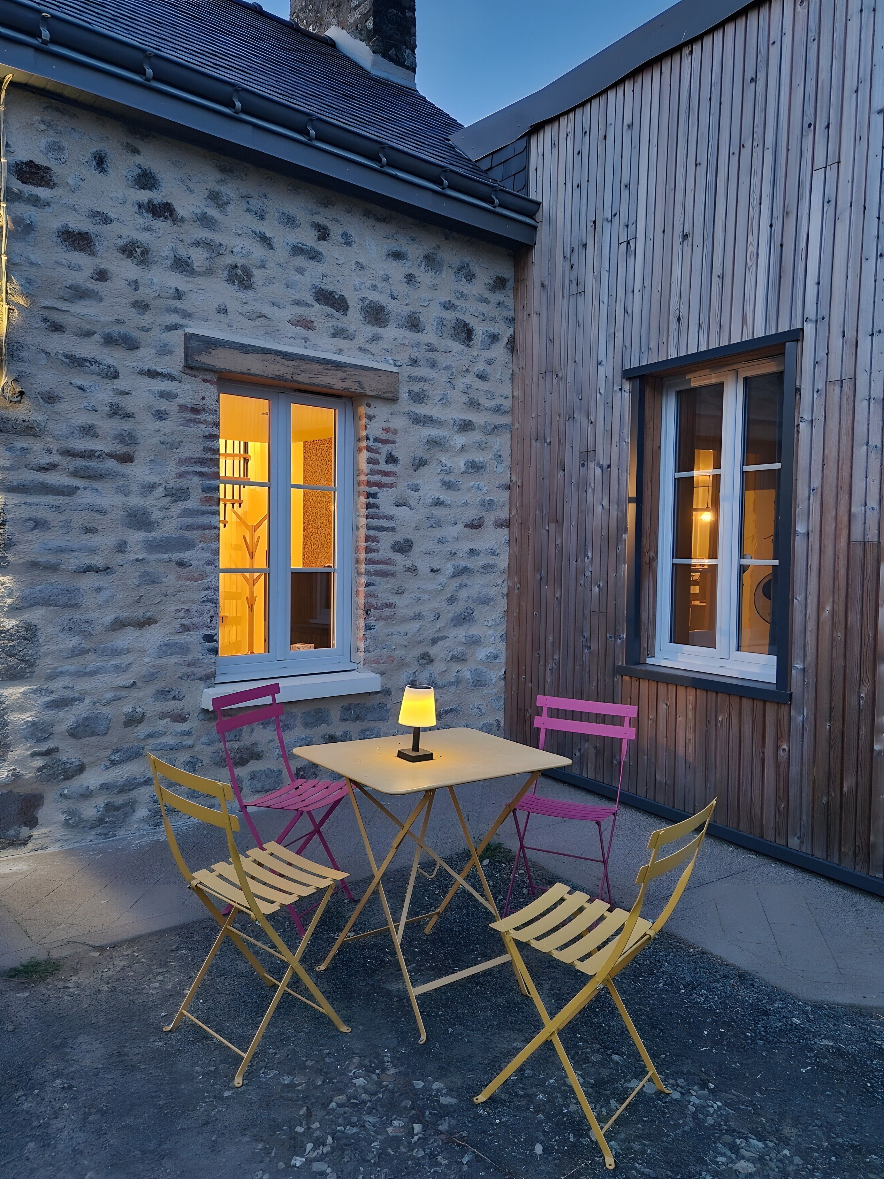 Un bâtiment en pierre avec une extension en bois, une table avec des chaises et une lampe.