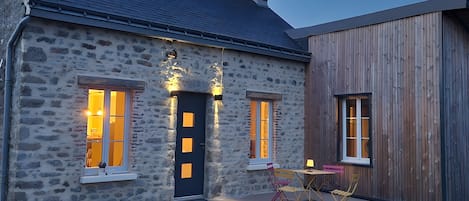 Une maison en pierre avec une porte sombre, deux fenêtres illuminées, une petite table avec des chaises et une extension en bois.