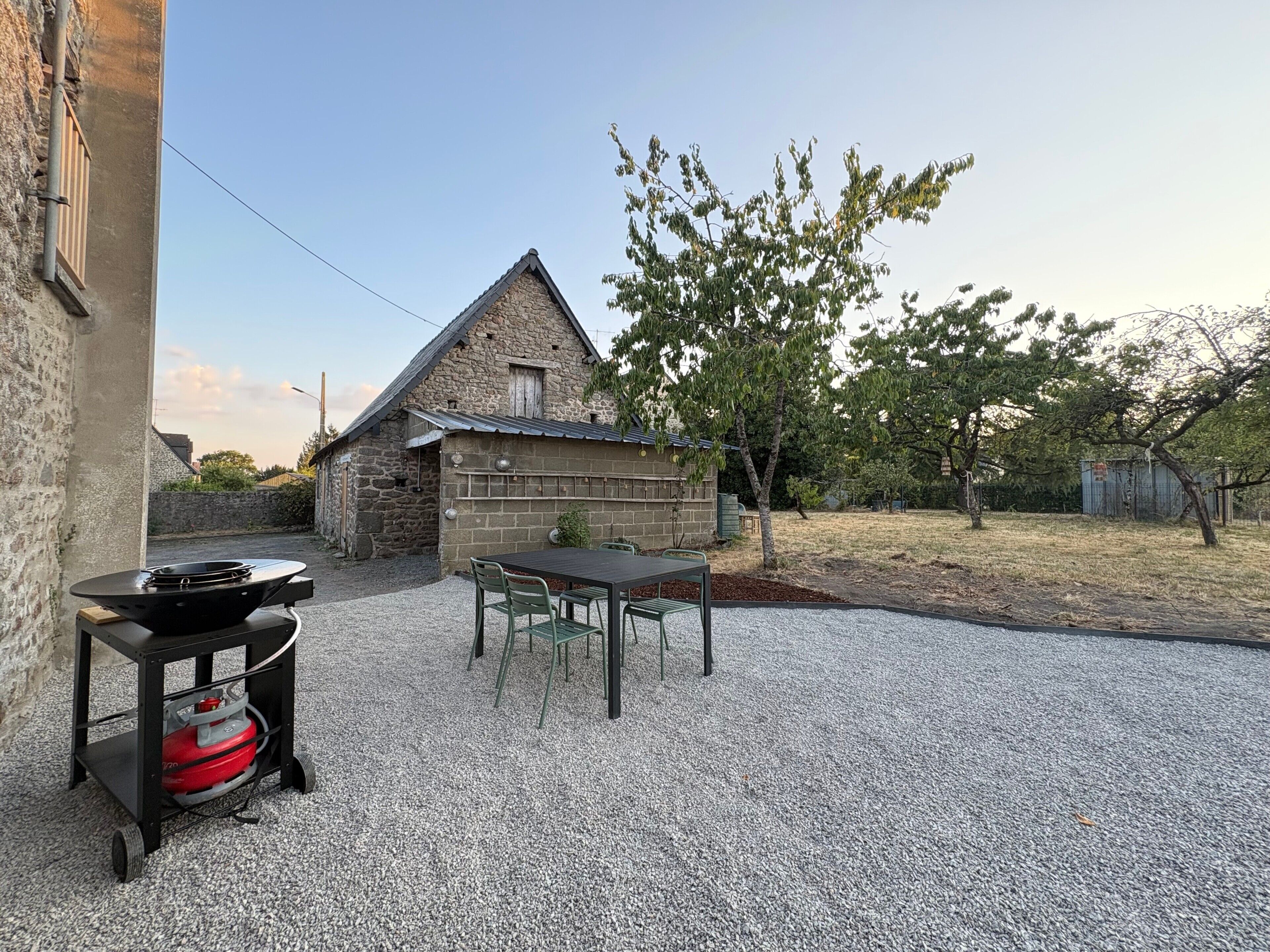 Een gebouw van steen met een terras van grind, een tafel met stoelen en een gasfles.