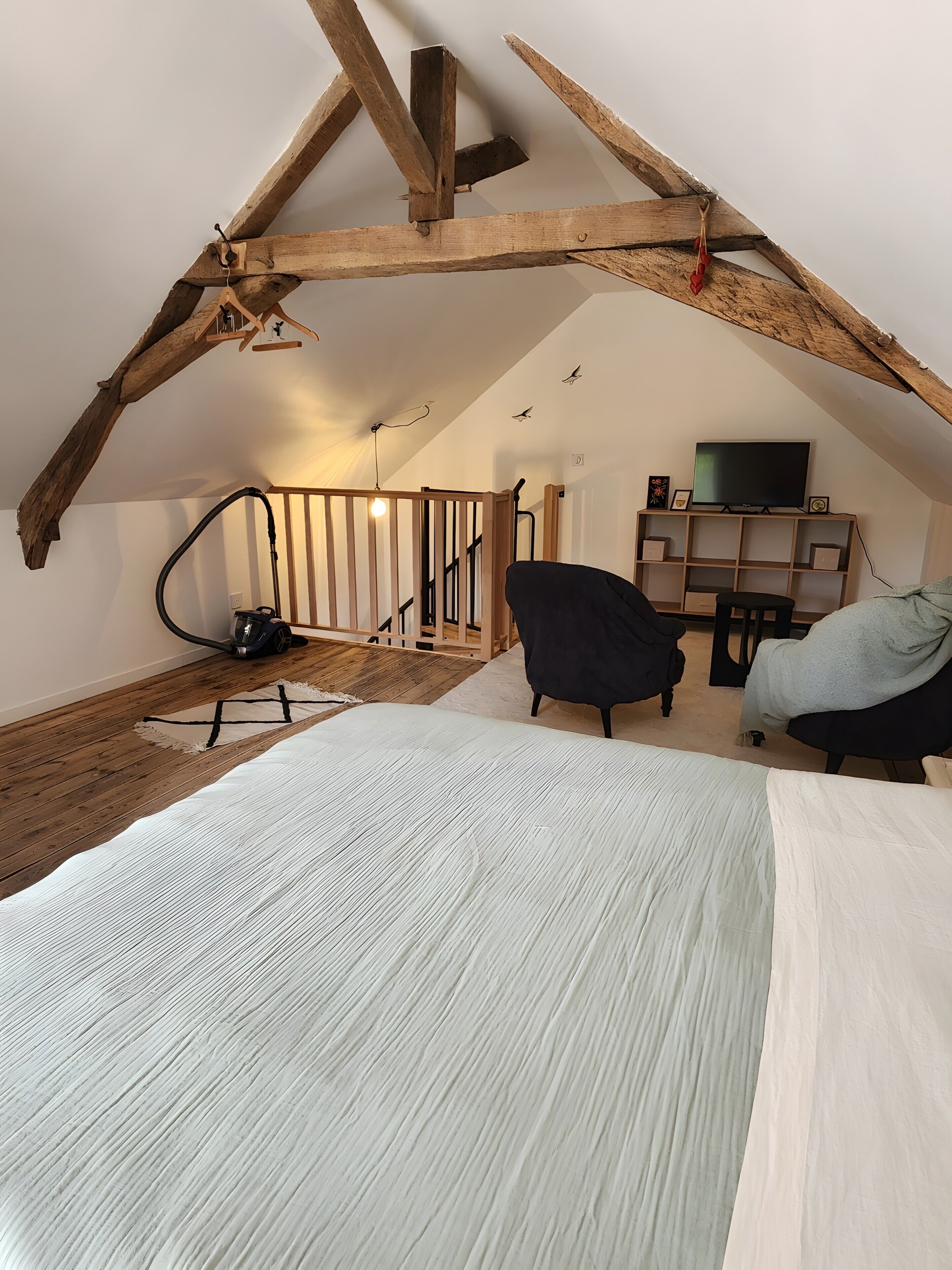 Une chambre à coucher aménagée dans un espace mansardé, avec des poutres apparentes, un lit, une chaise, un bureau, un aspirateur et un téléviseur posé sur une étagère.