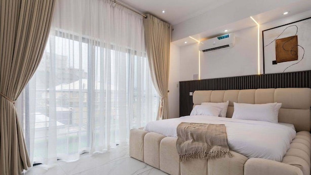 Un dormitorio moderno con una cama grande, una ventana con cortinas traslúcidas y un equipo de aire acondicionado instalado en la pared.