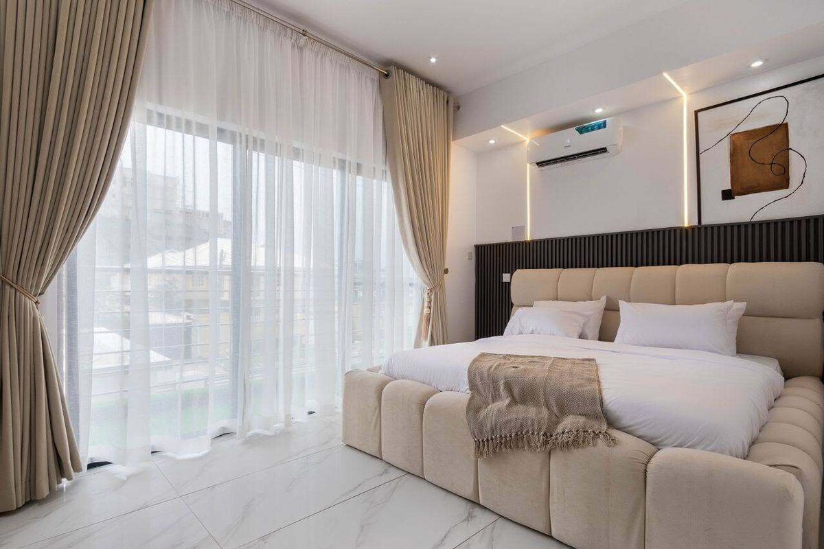 A modern bedroom with a large bed, a window with sheer curtains, and a wall-mounted air conditioning unit.