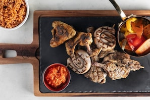 Uma refeição com carnes grelhadas, legumes e arroz, servida em uma lousa.