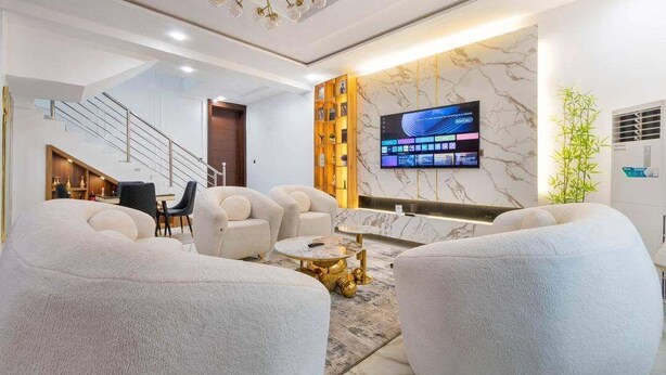 A modern living room with a flat-screen TV, plush seating, a staircase, and a decorative wall. - Shu Garden Hotel (Lagos)