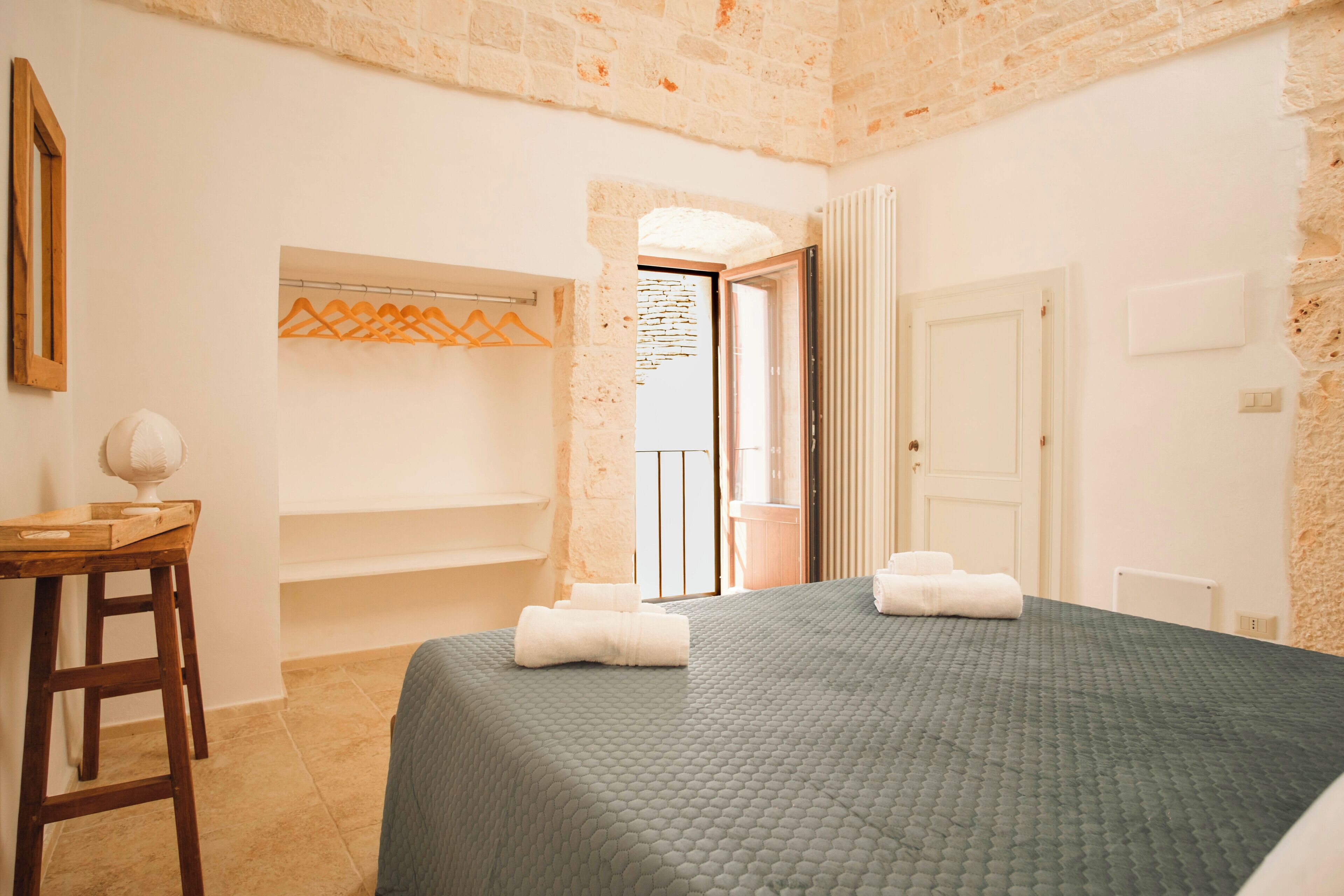 Une chambre d’hôtel avec un lit, un tabouret en bois, un porte-serviettes et une porte donnant sur un balcon.