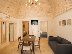 A room with a brick wall, a dining table with chairs, a sofa, and a television mounted on the wall. - Stone House—A Stone House in the Heart of Alberobello (Alberobello)