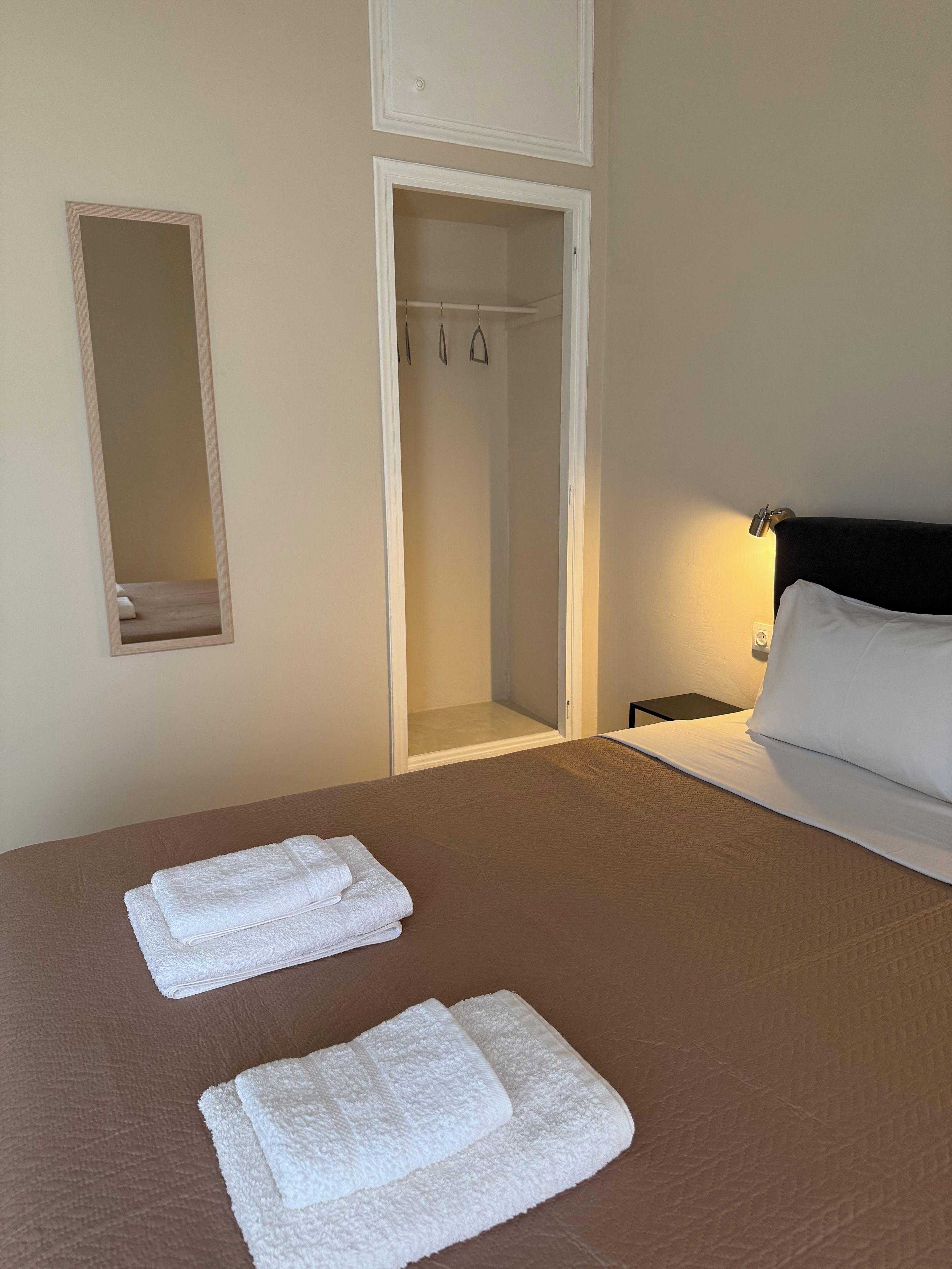 Una camera d'albergo con un letto, due asciugamani piegati con cura, un comodino con una lampada e un armadio con uno specchio.