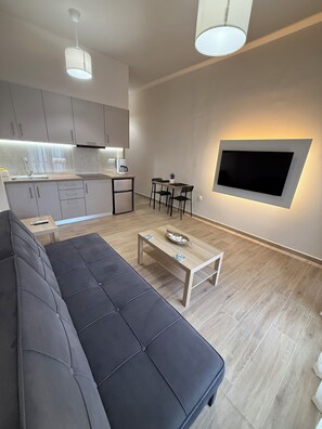 Un espai de viure modern amb un sofà, una taula de centre, un televisor fixat a la paret i una zona de cuina amb armaris i una superfície de treball.