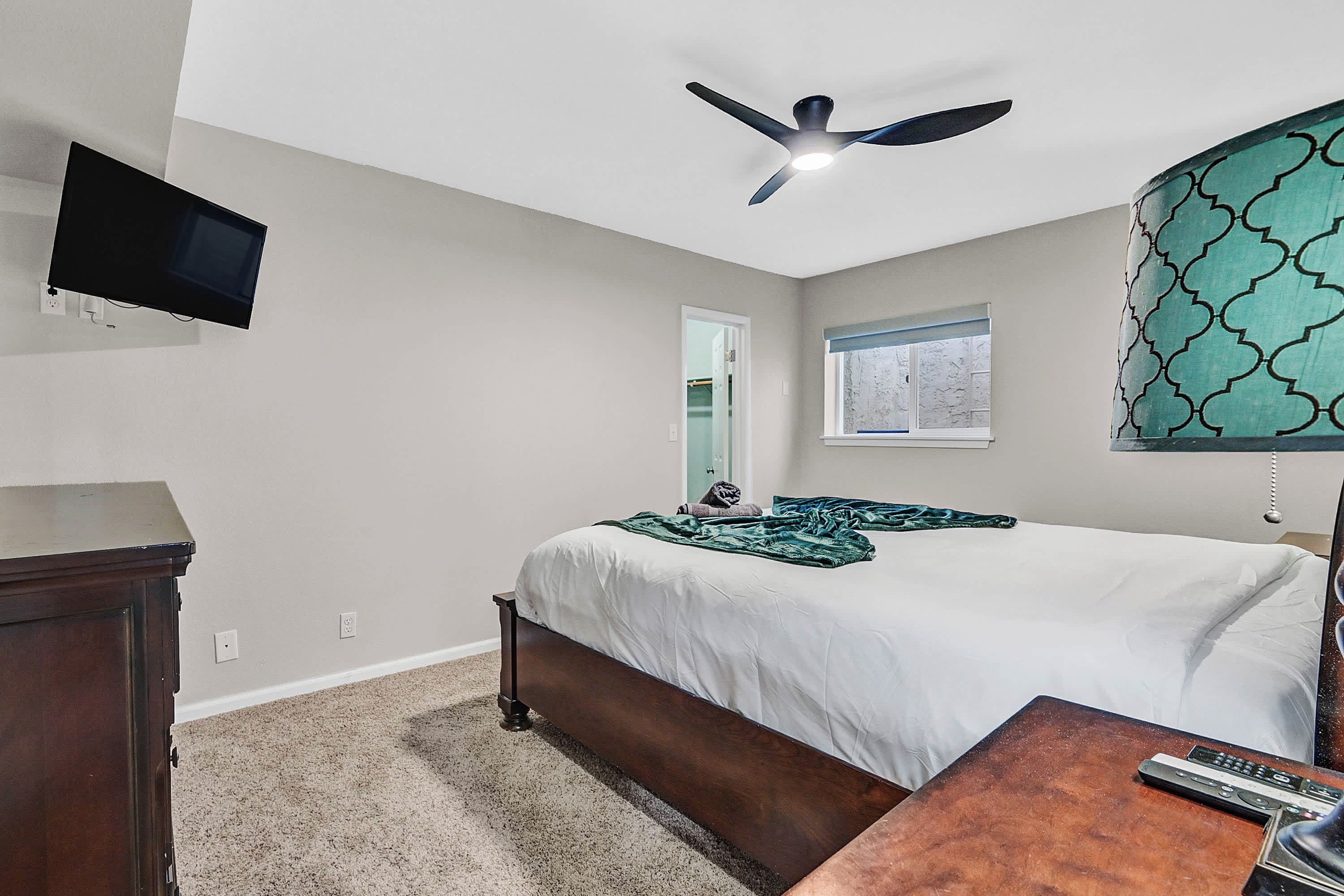 A bedroom with a bed, a desk, a chair, a ceiling fan, a TV mounted on the wall, and a window with a view.