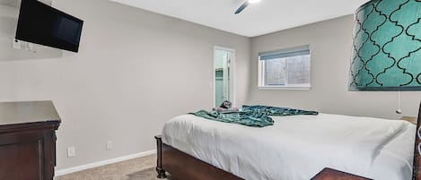 A bedroom with a bed, a desk, a chair, a ceiling fan, a TV mounted on the wall, and a window with a view.