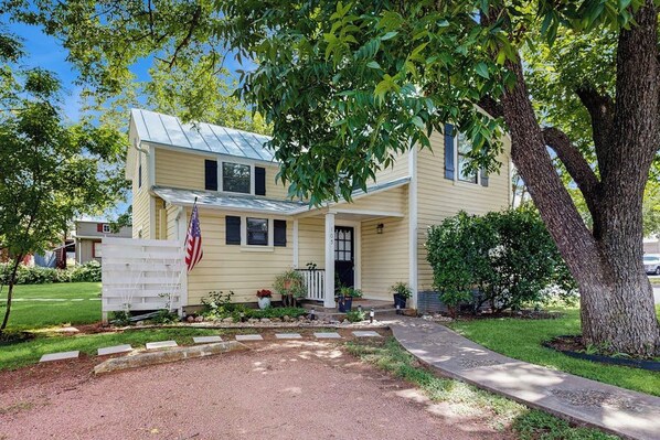 A two-story house with a front porch, a flag, and a large tree in the yard. - Beautiful Two-level Farmhouse w/ Garden Views, Free Wifi, & Outdoor Dining Area (Fredericksburg)