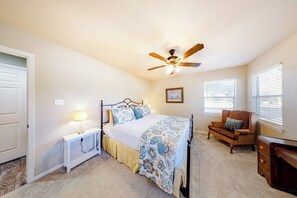 Ein Schlafzimmer mit Bett, Nachttisch, Lampe, Deckenventilator, Fenster und einem Sessel.