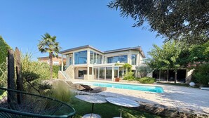 A modern two-story house with a swimming pool, surrounded by lush greenery and outdoor furniture.