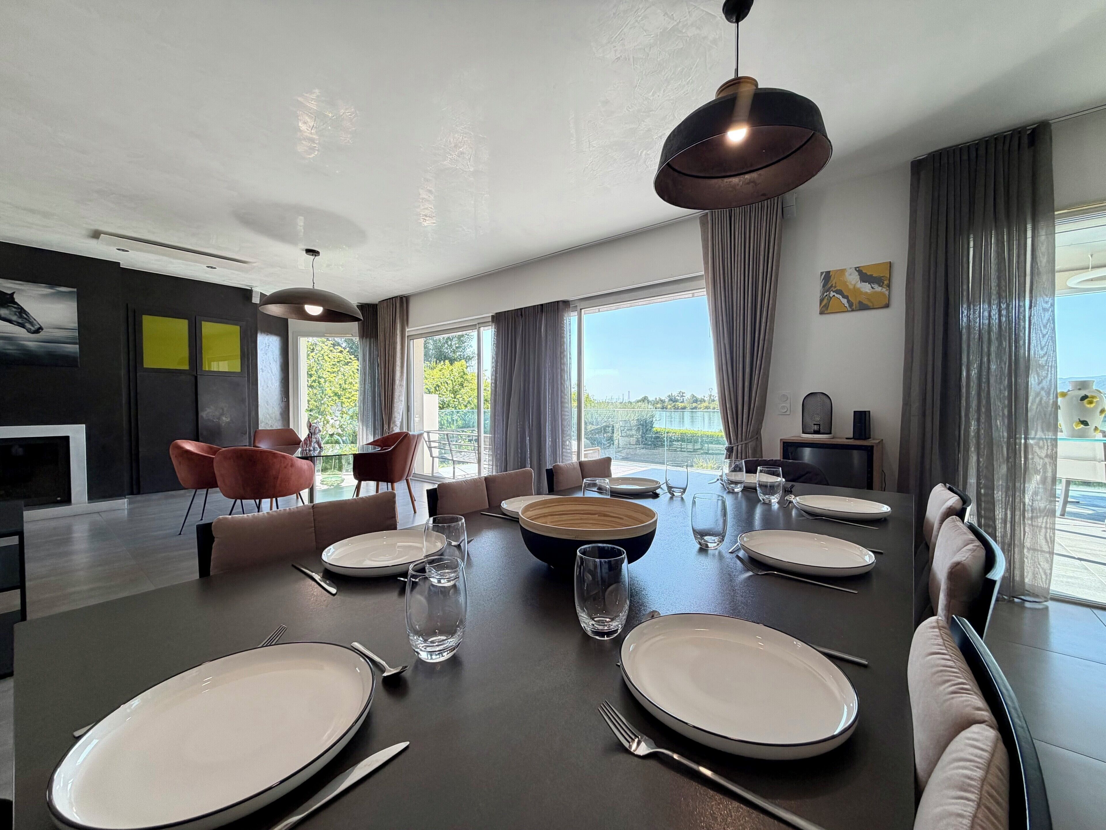 Un espace de repas moderne, avec une grande table dressée pour six personnes, entourée de chaises, et une vue sur l’extérieur grâce à de grandes fenêtres.