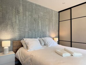 A modern hotel room with a large bed, white linens, a nightstand with a lamp, and a textured wall.