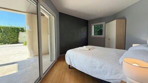 A modern bedroom with a large bed, a nightstand, a wardrobe, and a window with a view of greenery.