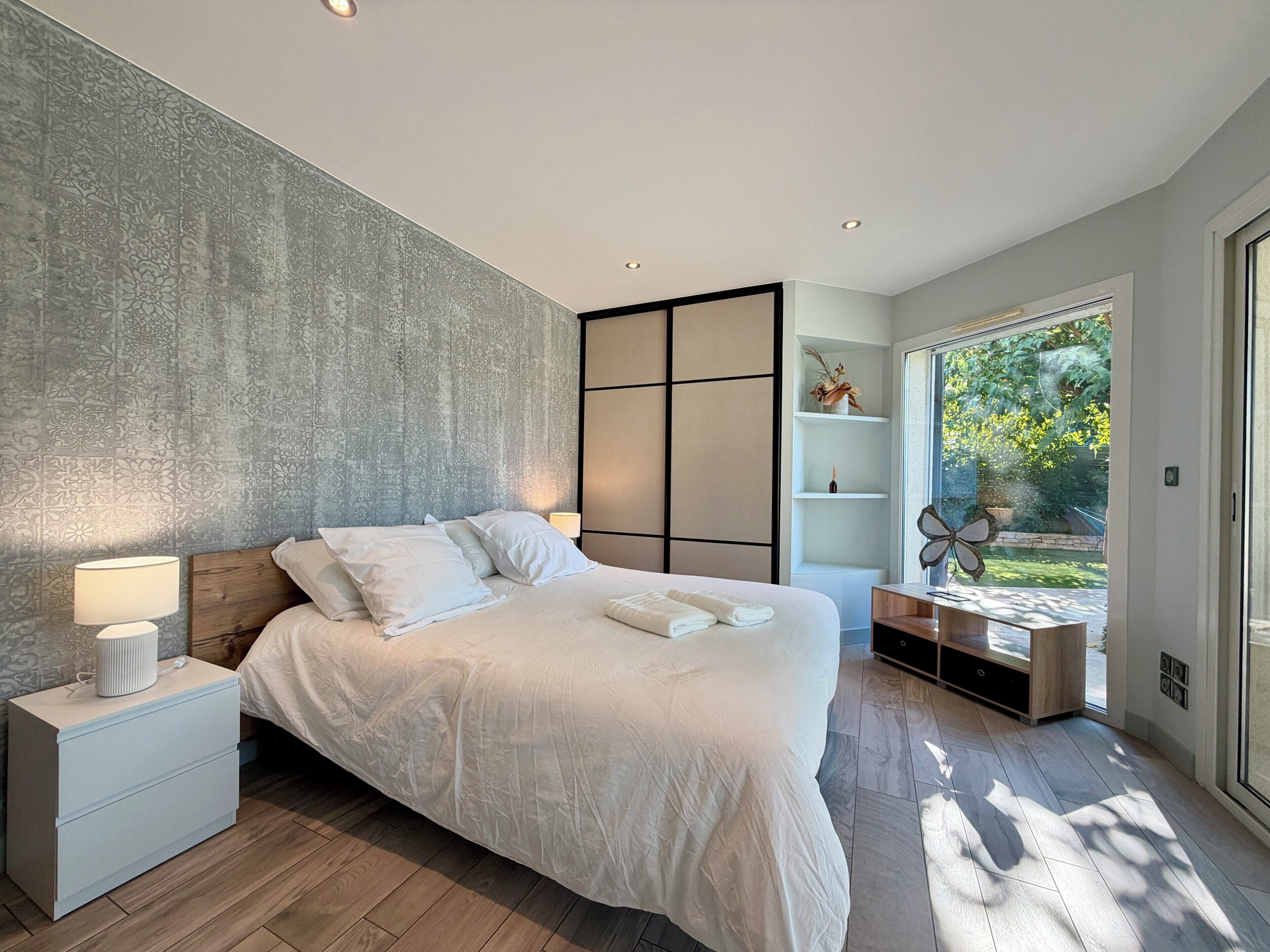 Een moderne slaapkamer met een groot bed, nachtkastjes, een wandkast en uitzicht op groen via een glazen deur.