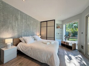 Une chambre moderne avec un grand lit, des tables de chevet, une commode, une armoire intégrée et une vue sur la verdure grâce à une porte vitrée.